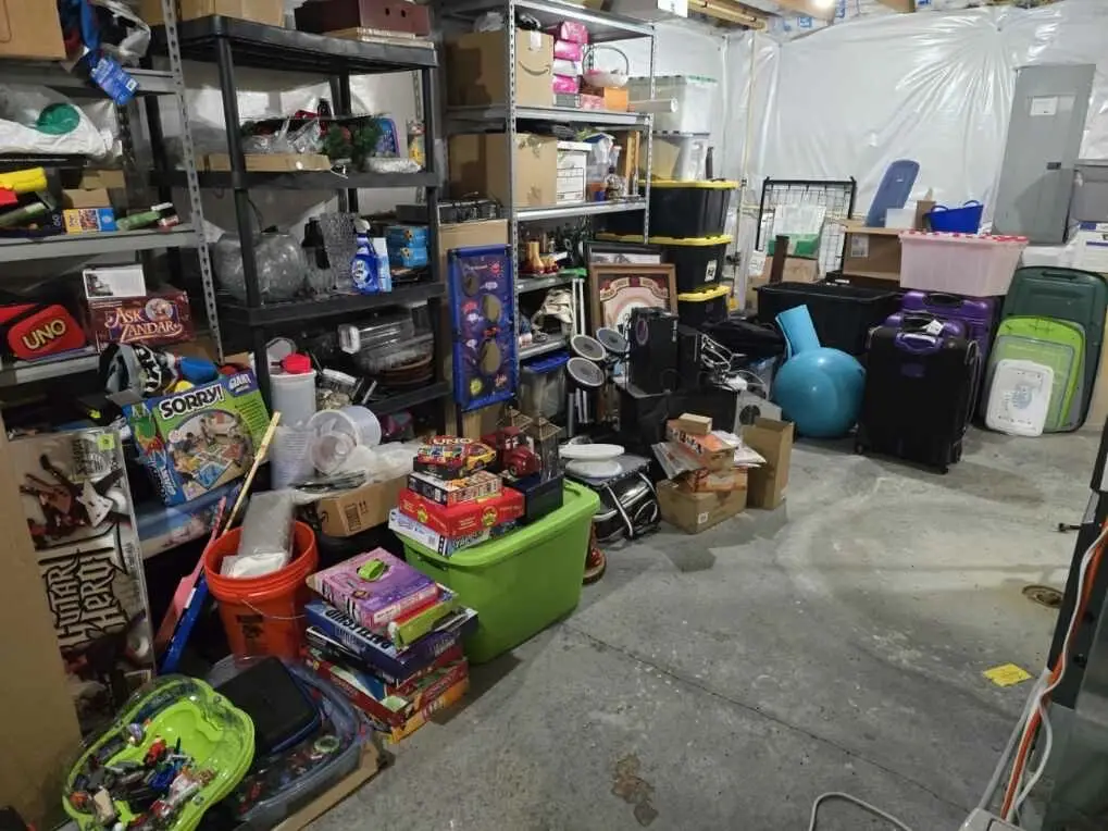 Before — board games, a green tote, an orange bucket and suitcases spread across the concrete floor in front of full shelving, basement in Union, KY