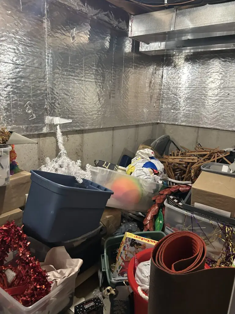 Before — a tinsel tree, a blue tub, garlands and wooden shelf parts piled against the foil insulation, basement storage room in Union, KY