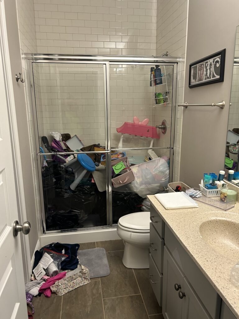 Before — a walk-in shower filled to the glass with black bags, toys and boxes, with clothes on the floor, guest bathroom in Union, KY