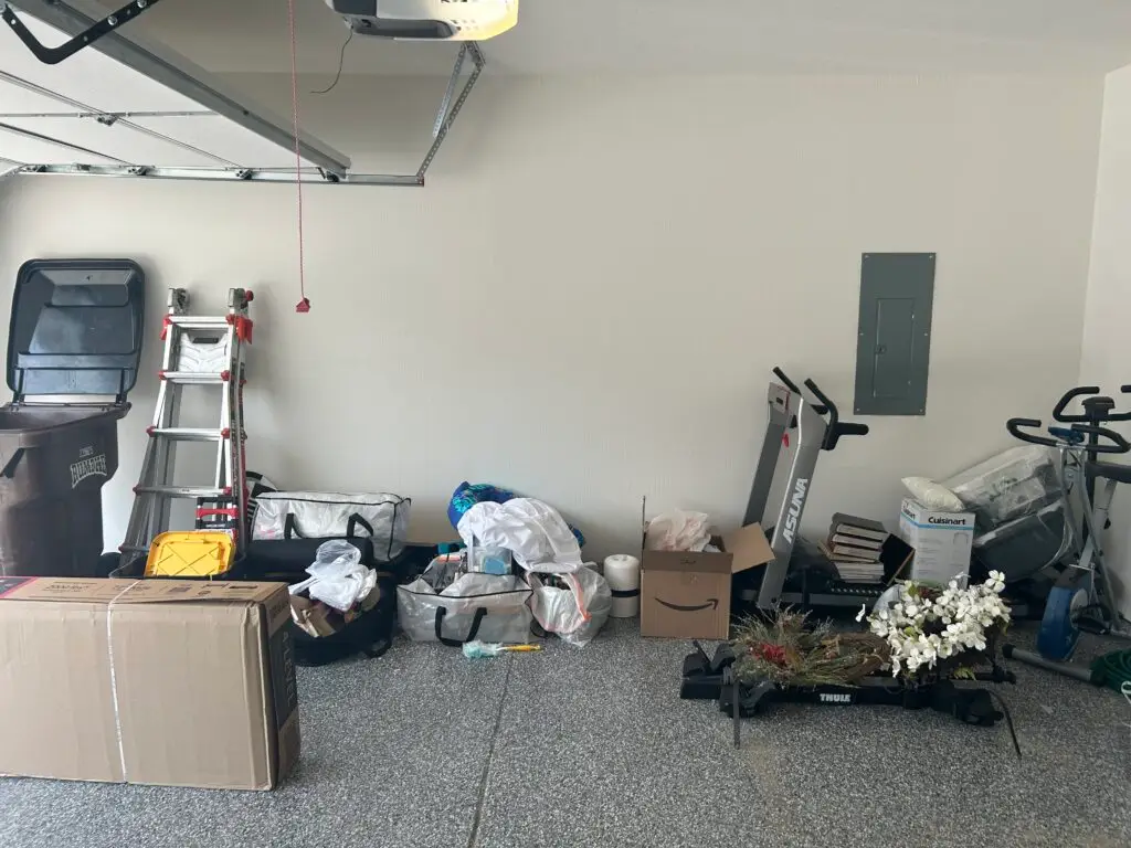Before — a treadmill, an exercise bike, a ladder, duffel bags and boxes lined along the wall of a garage with an epoxy floor, garage in Union, KY