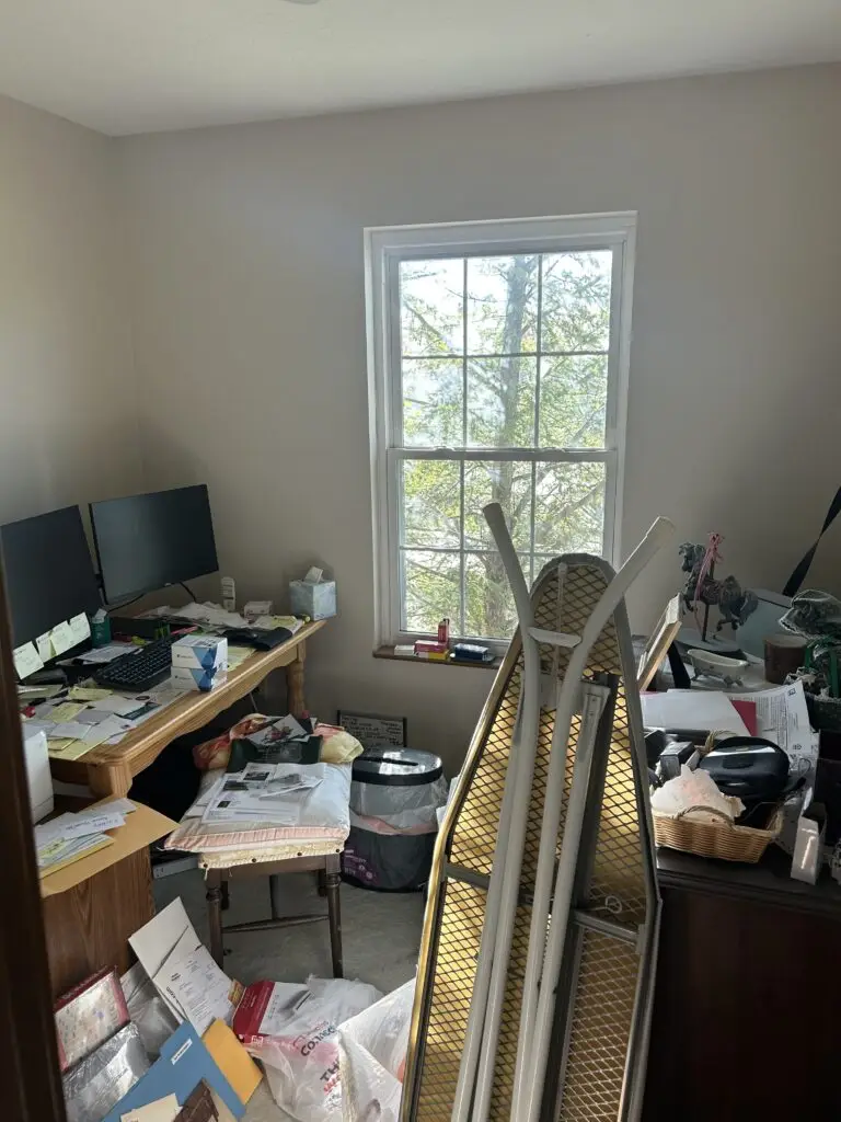 Before — a desk buried under paperwork with an ironing board and shopping bags across the floor, home office in Elsmere, KY