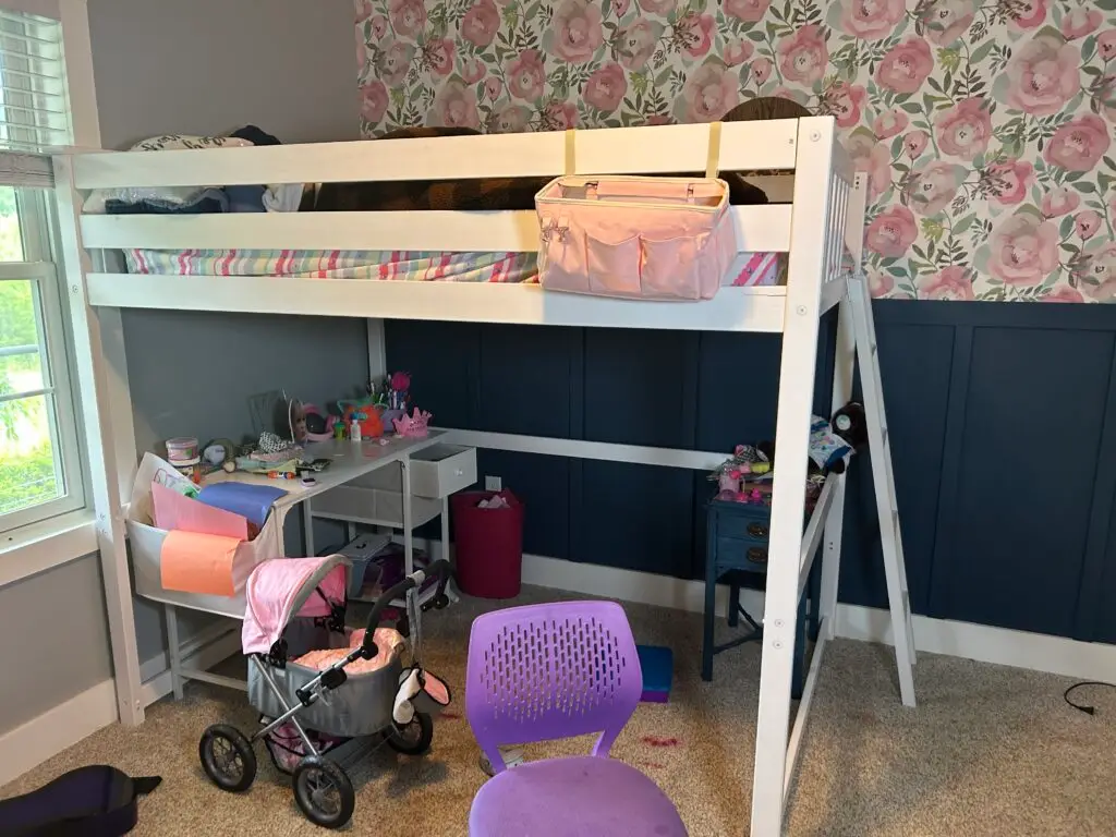 Before — a cluttered desk under the loft bed with toys and a doll pram across the carpet, girl's bedroom in Covington, KY