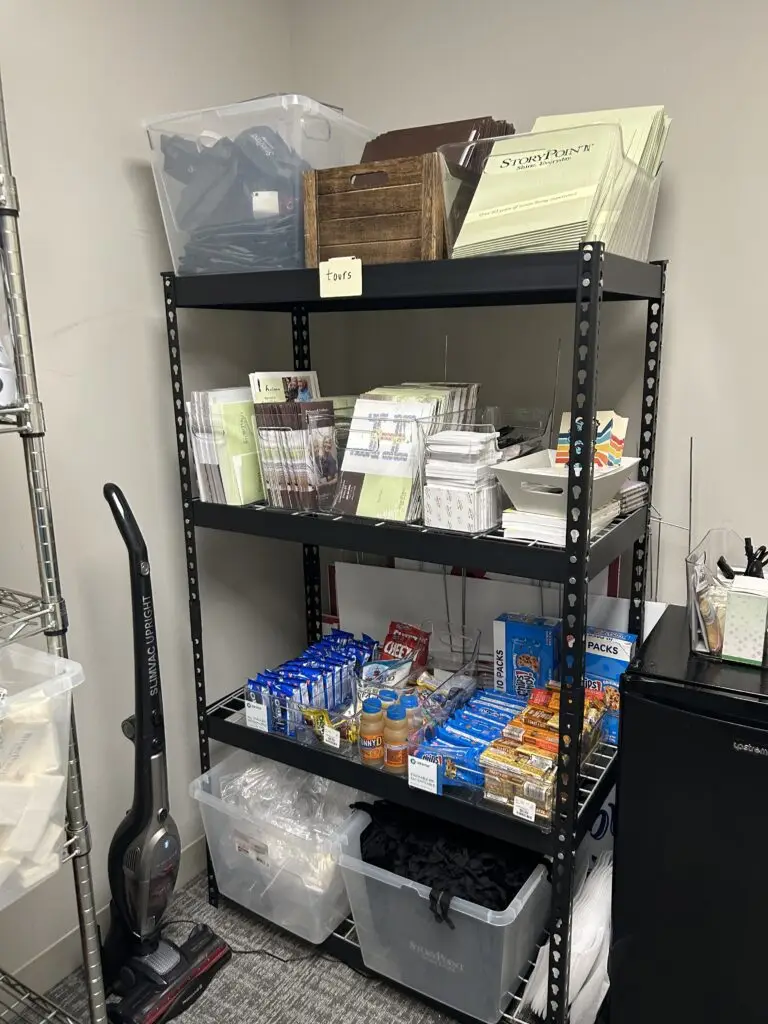 After — black shelving labeled by use with tour folders on top, brochures in acrylic holders and snacks in labeled trays, senior living storage room in Union, KY