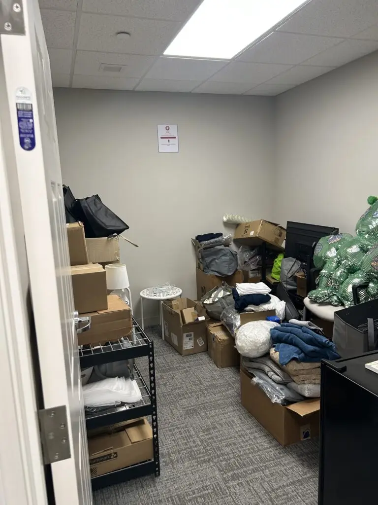 Before — cardboard boxes, folded linens and mesh bags piled across the floor beside a half-used wire rack, senior living storage room in Union, KY